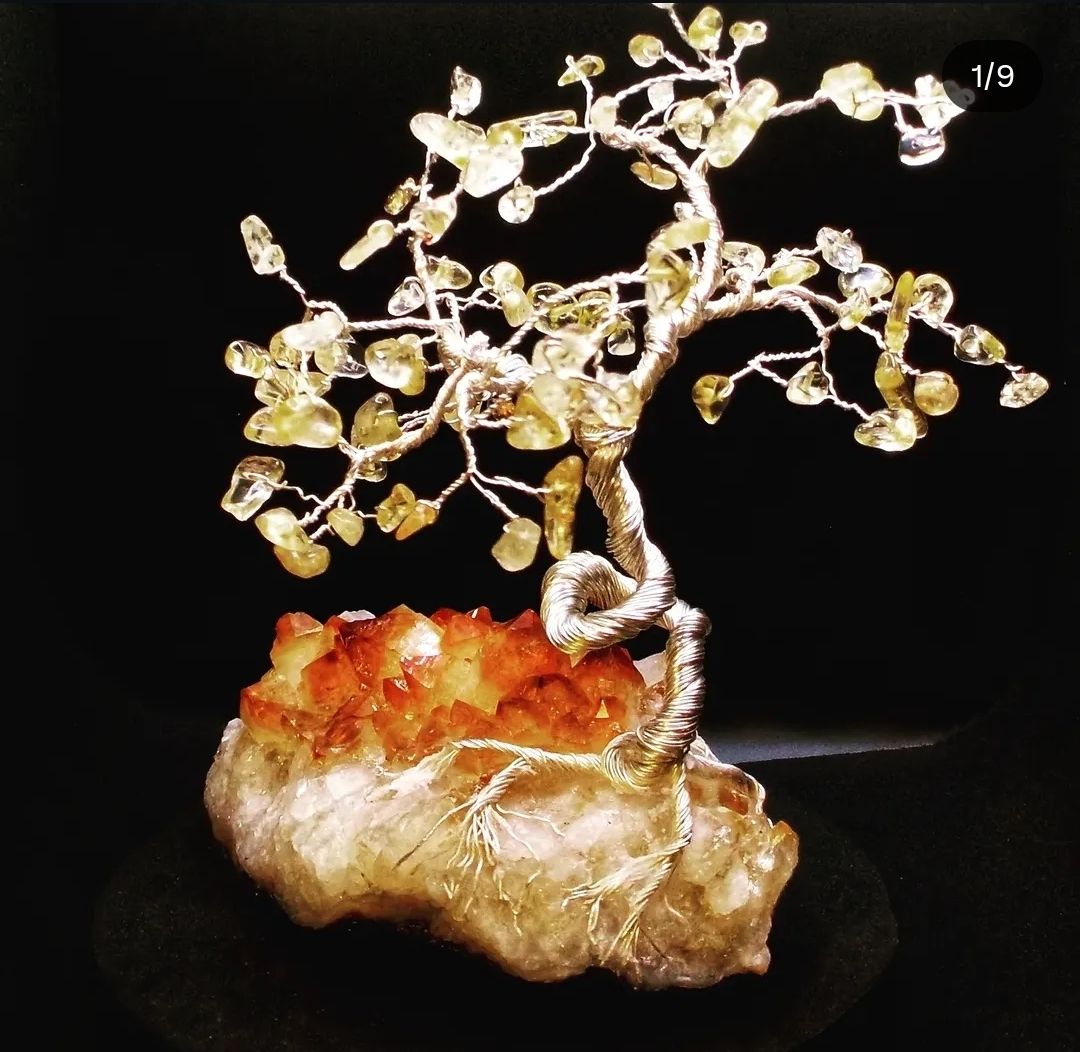 A decorative wire tree sculpture with yellow gemstone leaves, perched on a cluster of reddish-orange crystals, set against a black background.