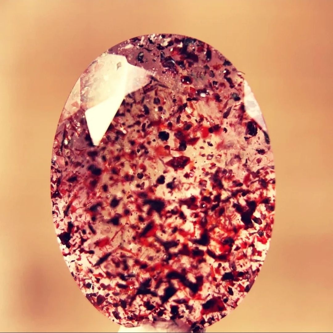 Close-up of a translucent pink gemstone with intricate patterns and dark speckles, displayed against a soft, blurred background.