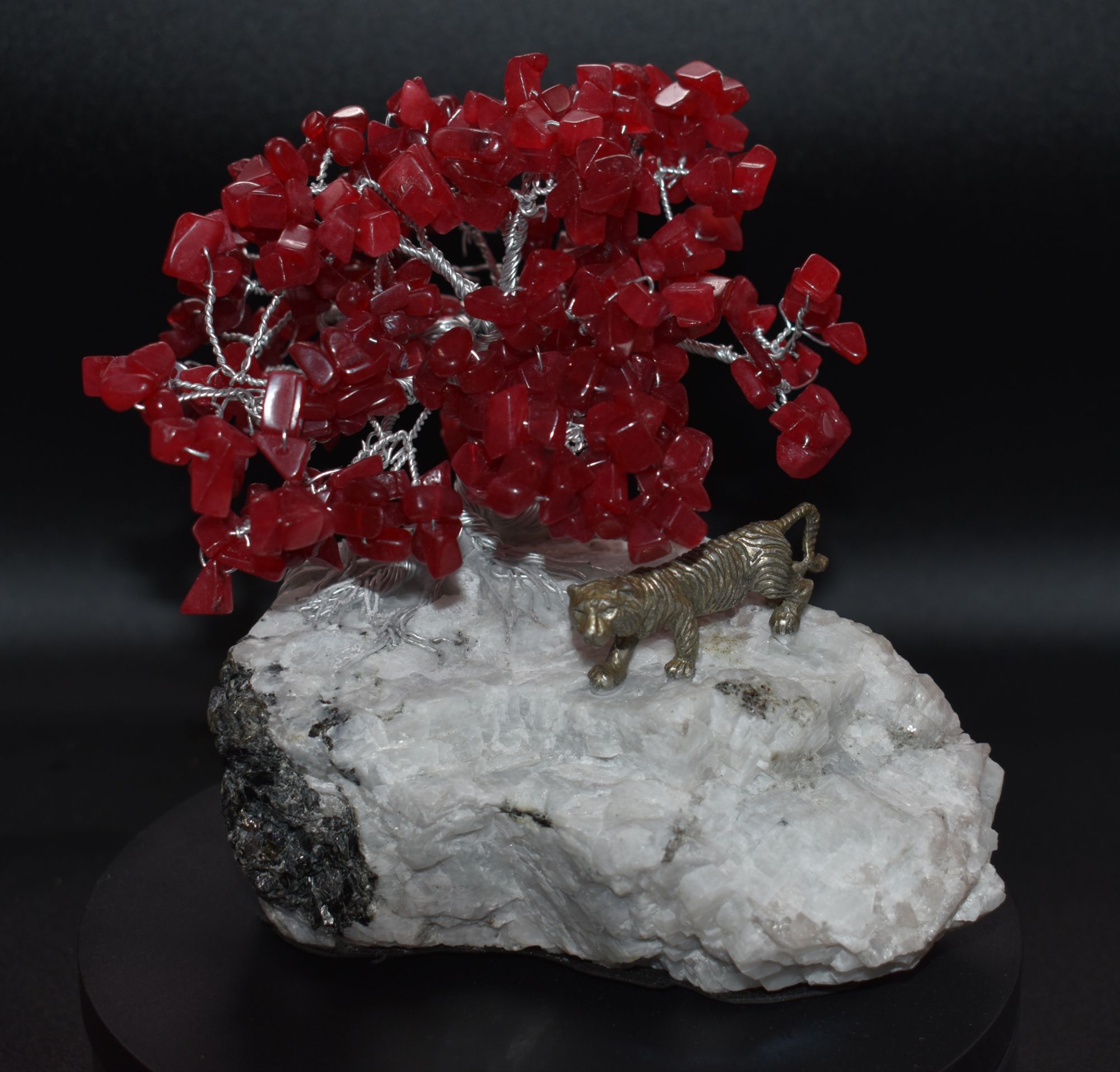 A decorative sculpture featuring a tree made of red crystals with a silver wire trunk and branches, positioned on a white and black rock base, alongside a small metal tiger figure.