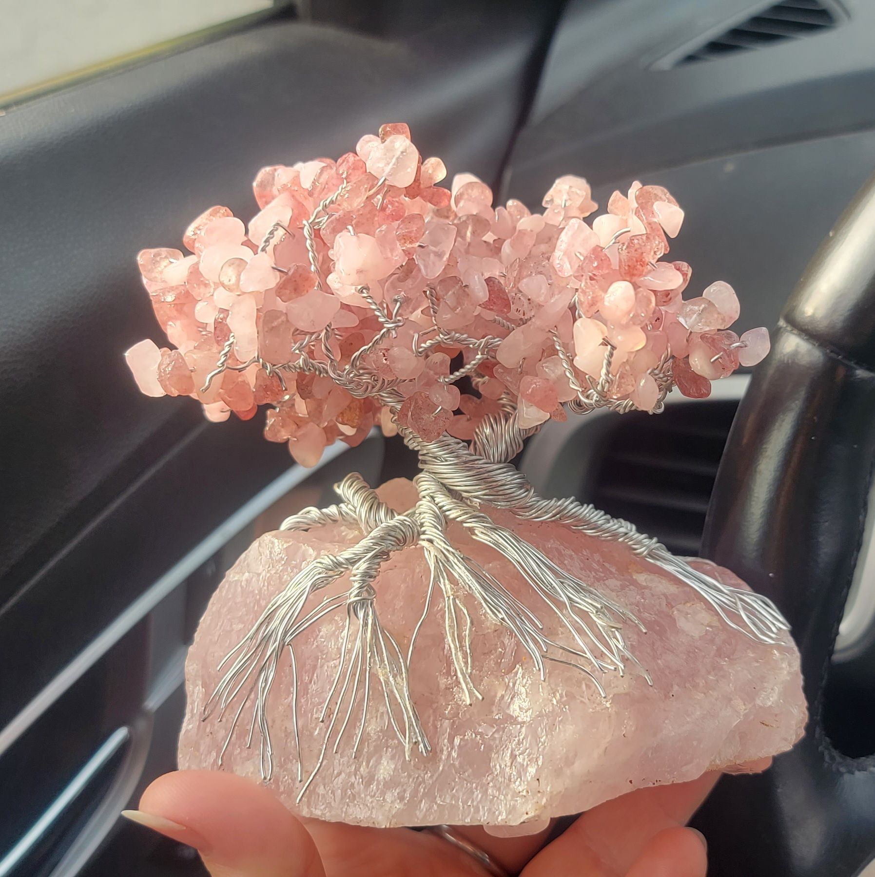 A handcrafted crystal tree sculpture made of rose quartz chips, with a twisted wire trunk and roots, resting on a large piece of rose quartz, held in a person's hand.
